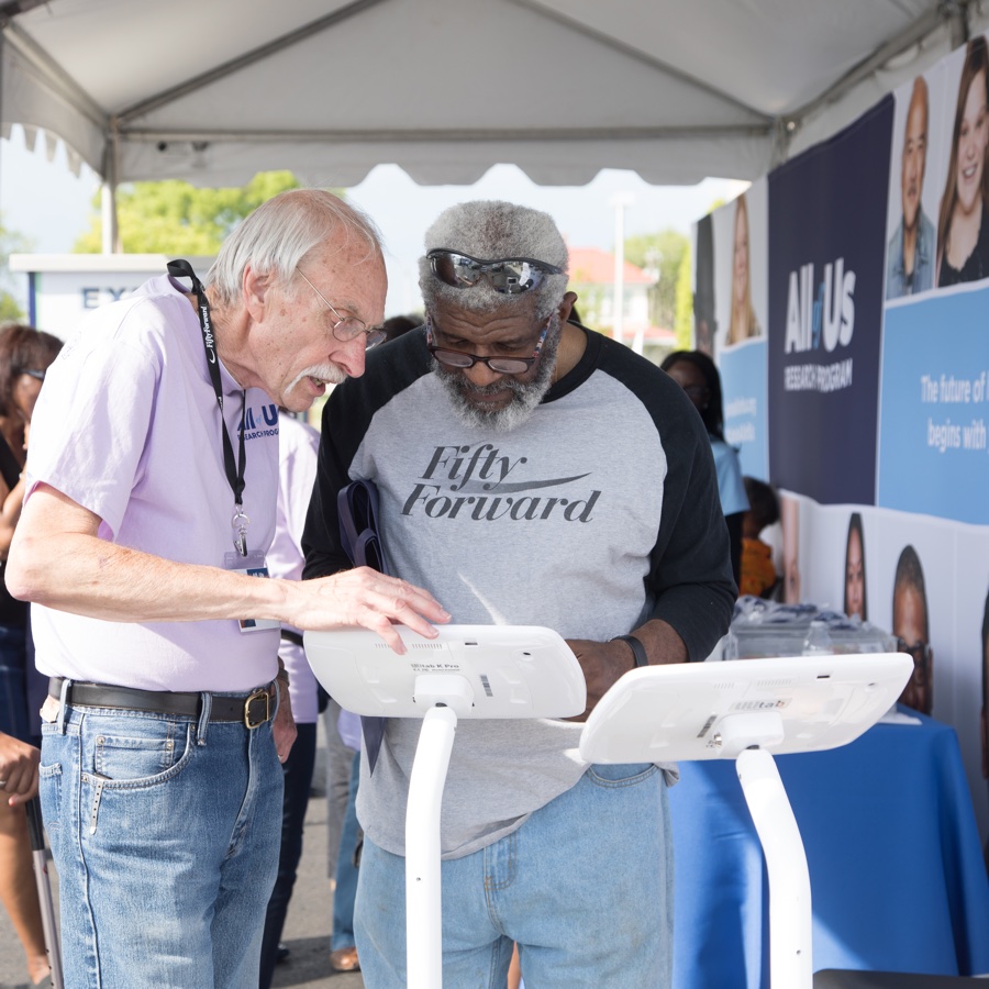 Two senior-aged men review All of Us Research Program information on a kiosk at an outdoor event.