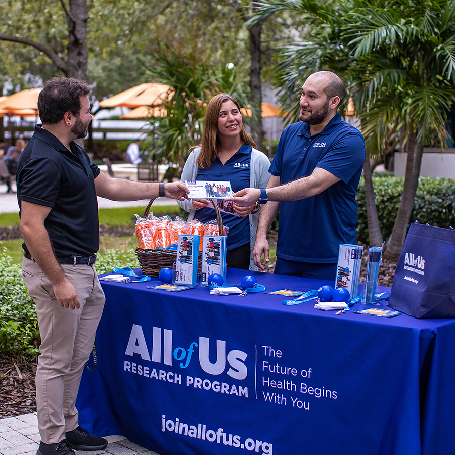 A woman and a man, dressed in All of Us Research Program shirts, stand behind an information table covered in All of Us communications materials, while engaging with a man and handing him a flyer about the program.