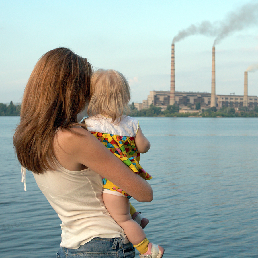 other and child look out across the water at a large industrial plant spewing emissions