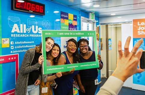  Four women pose for a photo behind All of Us branded selfie frame.