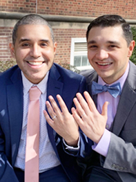 Two men sitting side by side, smiling as they hold up their hands to show off their wedding rings.